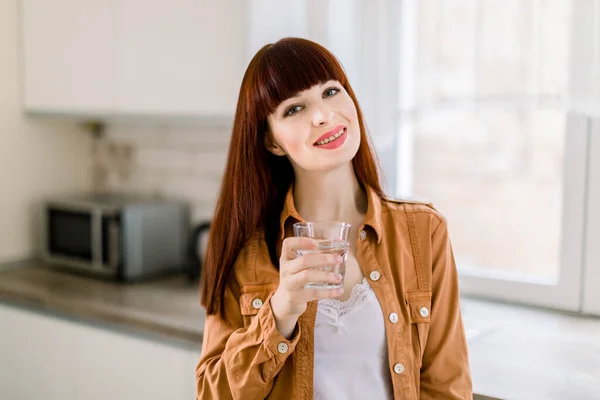 Encantadora joven sonriente con camisa de mostaza casual, bebiendo agua mientras está de pie en la cocina y mirando a la cámara. Estilo de vida saludable, concepto de dieta — Foto de Stock