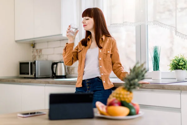 Joven mujer sonriente en ropa casual, bebiendo agua de minearl, mientras está de pie en la cocina moderna en casa, comenzando su día activo. Listo para desayunar sano y trabajar en línea — Foto de Stock