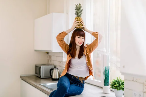 Foto de una hermosa mujer joven en jeans y camisa casual, sosteniendo la piña fresca en la cabeza y sonriendo a la cámara, posando en la cocina casera sentada en la encimera. Comida saludable, interior del hogar — Foto de Stock