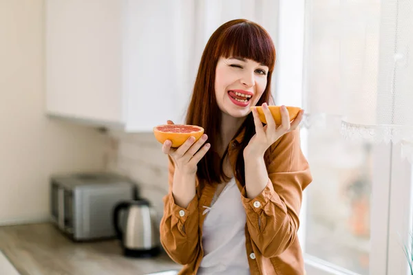 Foto horizontal de la divertida joven caucásica sentada en la encimera de la cocina y sosteniendo dos mitades de pomelo fresco, riendo ante la cámara mostrando su lengua. Menú diario saludable. Comida vegana — Foto de Stock