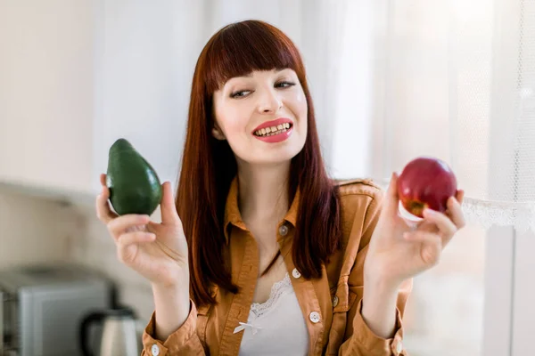 Egészséges evés, vegán kaja koncepció. Vonzó mosolygós fehér nő ül az asztalon a modern konyhában, kezében friss avokádó és vörös alma, néz az avokádó. Koncentrálj az arcra. — Stock Fotó