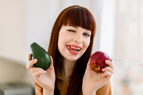 Крупним планом смішний портрет молодої жінки на кухні, що тримає свіже авокадо і червоне яблуко, сміється з камери, показуючи язик. Концепція здорового харчування та натуральної їжі — стокове фото