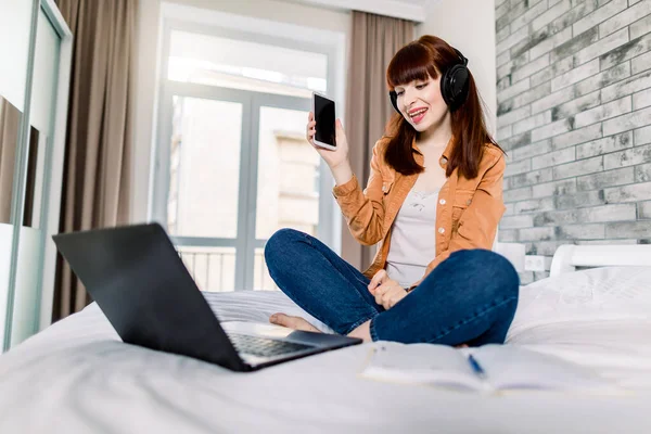 E-learning, estudar em casa. Foto de menina bonita em roupas domésticas casuais, usando fones de ouvido e celular enquanto trabalhava com laptop na cama no quarto elegante luz em casa — Fotografia de Stock