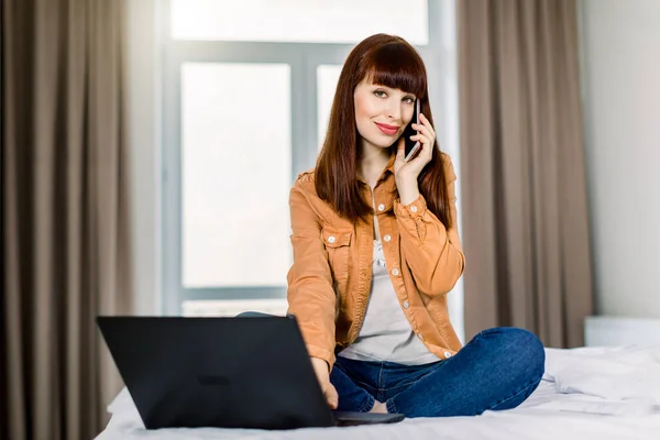 Jolie jeune femme d'affaires en tenue décontractée, assise sur le lit en pose de lotus, travaillant avec un ordinateur portable sur un lit, ayant un appel avec des collègues ou des clients. Concept de travail à domicile — Photo