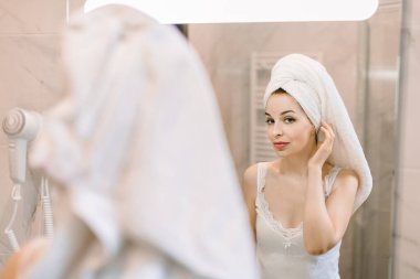 Spa kozmetik tedavi prosedürü, sabah hijyen konsepti. Kafası havluya sarılı genç, güzel bir kadın, aynadaki yansımaya bakıyor, duştan sonra banyoda.