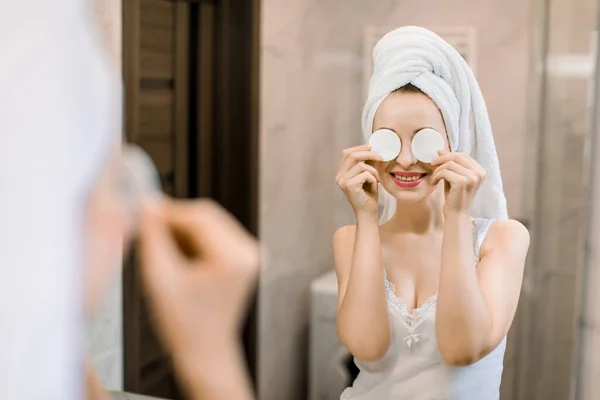 Fiatal, gyönyörű, mosolygós nő törülközővel a fején a tükörbe néz a fürdőszobában, nevet és szórakozik, miközben a szemét két pamut párnával takarja. Reggeli bőrápolás — Stock Fotó