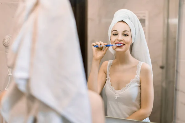 Refleksi di cermin tersenyum gadis muda dengan handuk mandi di kepala, menyikat gigi putih sehat dengan sikat gigi dan pasta gigi. Sehari-hari kebersihan mulut di kamar mandi di rumah — Stok Foto