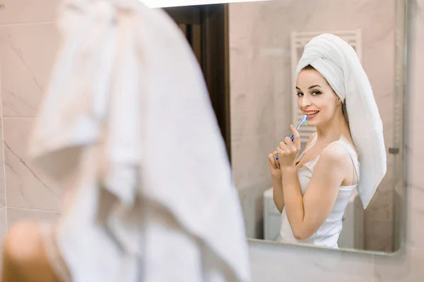 Uśmiechnięta biała młoda kobieta w ręczniku kąpielowym w głowie, patrząca w lustro w łazience, trzymająca szczoteczkę do zębów i sprawdzająca zęby. Poranna codzienna rutyna, leczenie jamy ustnej i higiena — Zdjęcie stockowe