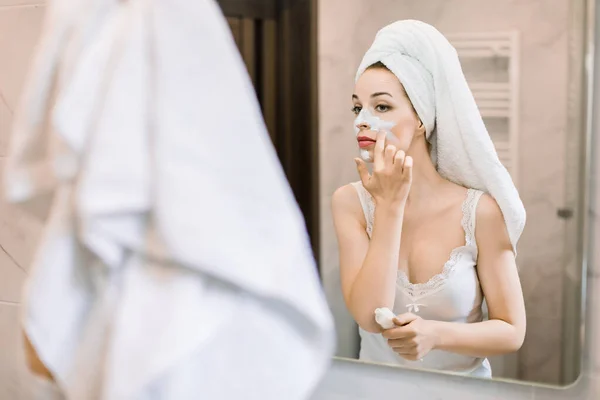 Lächelnde schöne Dame mit weißem Handtuch auf dem Kopf, die Hautpflege-Schlammmaske auf dem Gesicht, die in den Badezimmerspiegel schaut. Schönheitspflege am Morgen, Wellness-Behandlungskonzept — Stockfoto