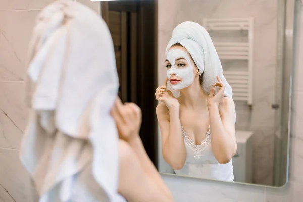 Blick aus dem Rückwinkel auf lächelnde attraktive junge Frau mit Handtuch auf dem Kopf, Anwendung Lehm Gesichtsmaske im Badezimmer. Hautpflege und Schönheitsbehandlung zu Hause — Stockfoto