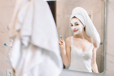 Ellerinde diş fırçası, yüzünde çamur maskesi olan banyoda eğlenen ve dans eden, kameraya bakan güzel beyaz kadının aynasında yansıması. Deri ve diş bakımı kavramı