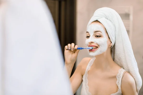 Mooie blanke dertiger jaren jonge vrouw met hoofd gewikkeld in een handdoek, met anti-aging gezichtsmasker op de huid, kijkend in de badkamer spiegel terwijl ze haar tanden poetst met tandenborstel. Ochtendroutine concept — Stockfoto