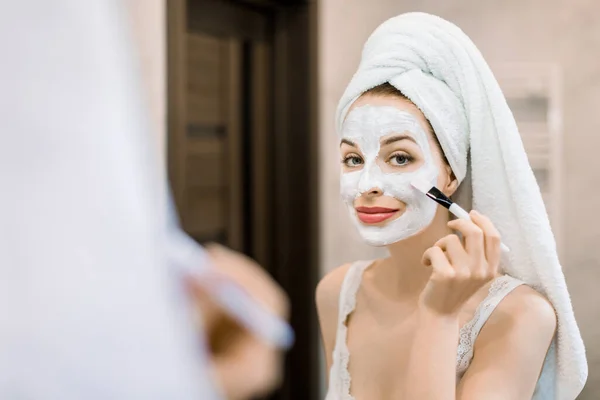 Ομορφιά φροντίδα του δέρματος, καλλυντικά και την έννοια της υγιεινής. Νεαρή γυναίκα με πετσέτα στο κεφάλι, κοιτάζει στον καθρέφτη, κρατώντας πινέλο και απλώνοντας μάσκα λάσπης προσώπου στο πρόσωπό της στο μπάνιο — Φωτογραφία Αρχείου