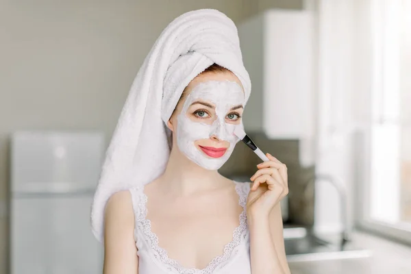 Ładna młoda, kaukaska kobieta z włosami owiniętymi w ręcznik kąpielowy, pozująca do kamery w nowoczesnej kuchni domowej, nakładając białą glinianą maskę błotną na twarz pędzlem kosmetycznym. Spa i piękno w domu — Zdjęcie stockowe