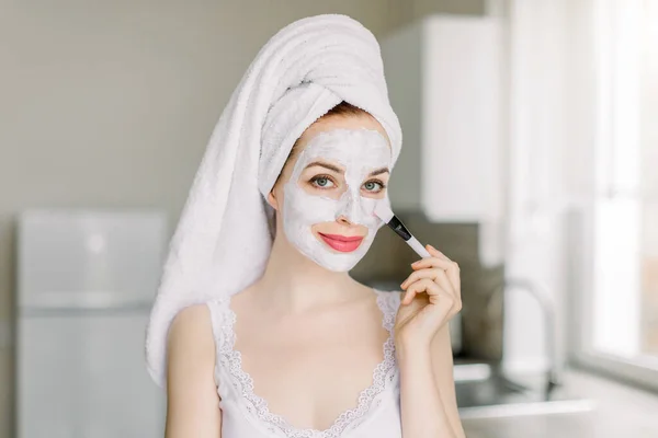 Mooie jonge blanke vrouw met haar gewikkeld in badhanddoek, poseren voor camera in de moderne huiskeuken, terwijl het aanbrengen van witte klei modder gezichtsmasker op haar gezicht met cosmetische borstel. Spa en schoonheid thuis — Stockfoto