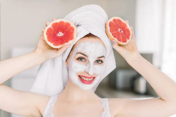 Jovem sorridente alegre após o chuveiro, com máscara de barro facial, posando para câmera em casa fundo interior com metades de toranja fresca. Conceito de cuidados com a pele e beleza em casa — Fotografia de Stock