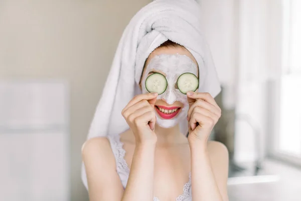 Muchacha caucásica joven divertida con máscara de arcilla para la piel de la cara y los pepinos en los ojos, posando en la cocina casera de luz y divertirse, disfrutando de procedimientos de belleza spa en casa —  Fotos de Stock