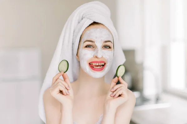 Mulher bonita rindo jovem com cabelo envolto em toalha de banho, com máscara facial em seu rosto segurando fatias de pepino fresco, sorrindo para a câmera. Cuidados com a pele, spa, beleza natural e conceito cosmetologia — Fotografia de Stock