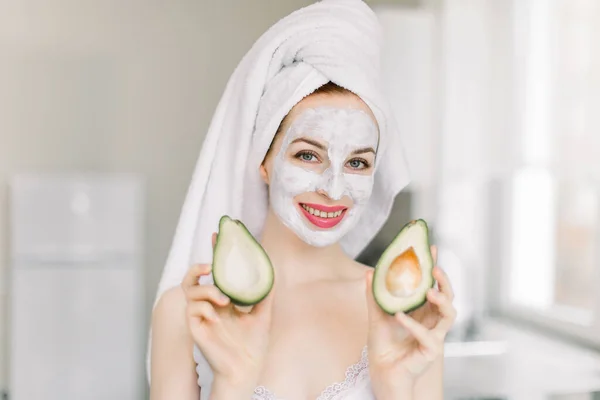 Portrait de charmante jeune femme enveloppée dans une serviette, debout à la maison cuisine et ayant journée spa avec masque facial, tenant avocats moitiés dans les mains et souriant à la caméra. Spa et concept de soins de la peau — Photo