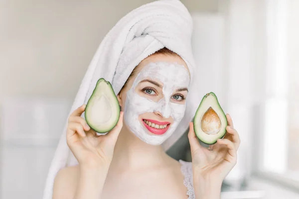 Beautiful young attractive girl with facial mask and hair wrapped in bath towel, holding halves of fresh avocado, posing to camera in home interior. Close up portrait. Spa, beauty and cosmetology — Stock Photo, Image