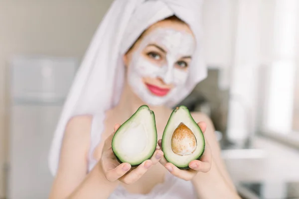 Young smiling attractive woman holding two halves of avocado, with anti aging facial cream mask, posing over blurred home kitchen background, showing avocado to camera. Focus on avocado — Stock Photo, Image