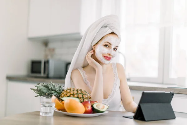 Glimlachende blanke jonge vrouw met witte anti-aging gezichtsmasker op gezicht, studeren, werken of online winkelen op tablet computer, terwijl ze aan de keukentafel met fruit thuis zit — Stockfoto