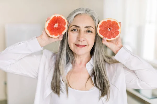 Beautiful senior woman in white shirt is looking at camera and smiling while standing with two halves of fresh grapefruit in light kitchen. Horizontal close up shot. Healthy eating concept — Stock Photo, Image