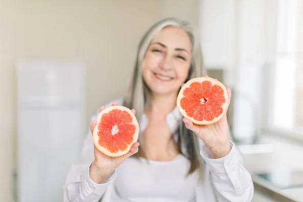 Mujer de mediana edad o mayor de pelo gris de pie en la cocina, sosteniendo mitades de pomelo fresco en las manos como promoción de una alimentación saludable y la prevención contra las enfermedades. Concéntrate en las manos con fruta —  Fotos de Stock