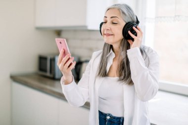 Müzikten zevk almak ve mutfakta eğlenmek. Neşeli yaşlı gri saçlı kadın kulaklıklardan müzik dinliyor ve gülümsüyor ya da en iyi arkadaşıyla video görüşmesi yapıyor. Yaşlılar ve teknolojiler