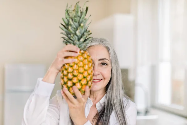 Middle age beautiful woman wearing white shirt, holding pineappe and hiding half of her face, looking at camera with happy smile while standing in modern kitchen at home. Healthy food — Stock Photo, Image