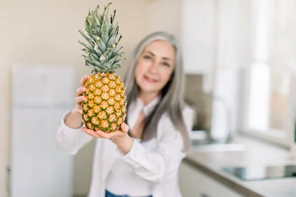 Healthy lifestyle, elderly people. Charming senior lady with long gray hair, standing in the modern kitchen at home and showing fresh pineapple to camera. Focus on pineapple — Stock Photo, Image