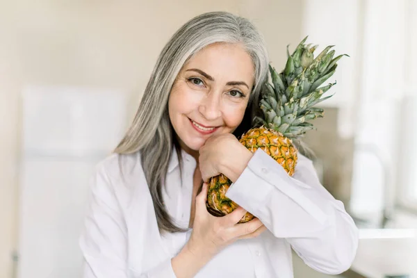 Close up horizontal beauty shot of happy senior gray haired woman holding fresh pineapple, looking at camera with smile, standing in modern light kitchen at home. People, portraits and healthy food — Stock Photo, Image