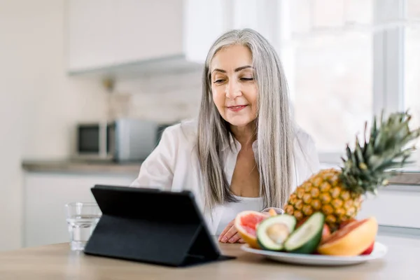 Όμορφη κομψή ηλικιωμένη γυναίκα με μακριά γκρίζα μαλλιά, κάθεται στο τραπέζι της κουζίνας με φρέσκα φρούτα και χρησιμοποιώντας ψηφιακή ταμπλέτα για τα κοινωνικά δίκτυα, εργάζονται σε απευθείας σύνδεση ή το μαγείρεμα — Φωτογραφία Αρχείου