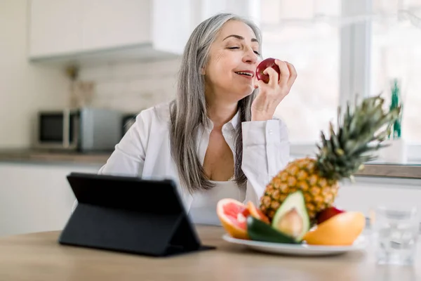 Όμορφη ηλικιωμένη συνταξιούχος με μακριά γκρίζα μαλλιά κάθεται στο τραπέζι της κουζίνας στο σπίτι και απολαμβάνει το μεσημεριανό της με φρέσκο μήλο και φρούτα. Υπολογιστής tablet στο τραπέζι — Φωτογραφία Αρχείου