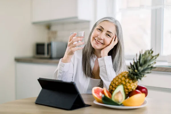 Beautiful retired gray haired woman with glass of water in kitchen, sitting at the table and looking at camera with nice smile, while having healthy fruit breakfast and working on tablet computer — Stock Photo, Image