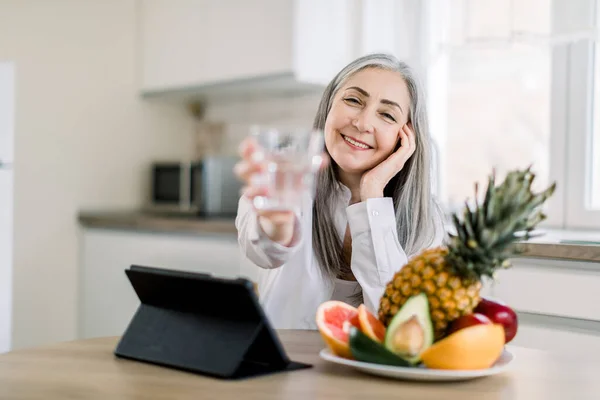 Joyful smiling Caucasian senior woman with long gray hair, showing glass of water to camera, while sitting at the table with fresh fruits and ipad in modern light kitchen. Focus on face — Stock Photo, Image