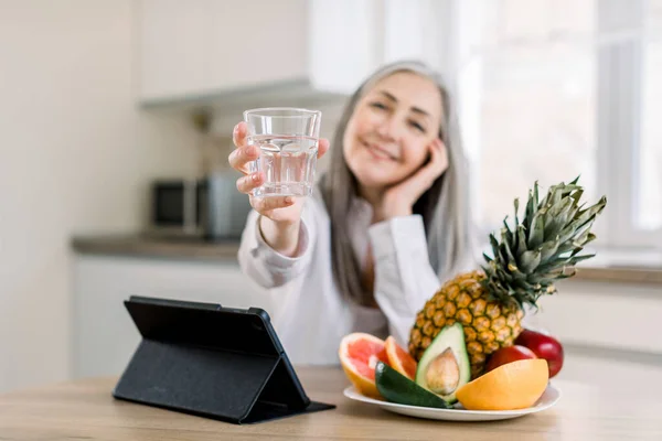 Joyful smiling Caucasian senior woman with long gray hair, showing glass of water to camera, while sitting at the table with fresh fruits and ipad in modern light kitchen. Focus on glass — Stock Photo, Image