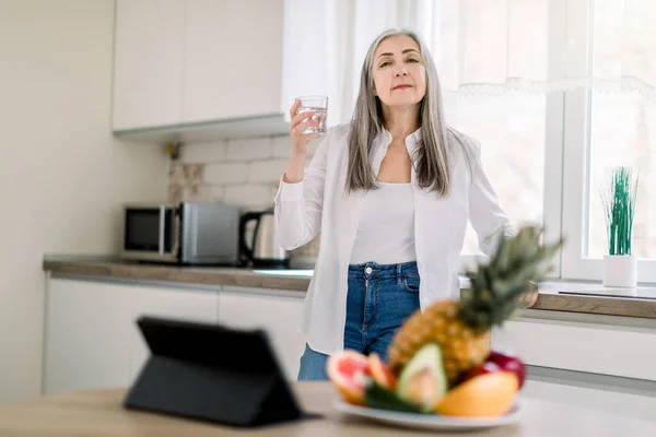 Healthy food for elderly people. Smiling senior woman in white shirt and jeans, drinking mineral water, while standing in modern kitchen at home, starting her active day, breakfast and work — Stock Photo, Image