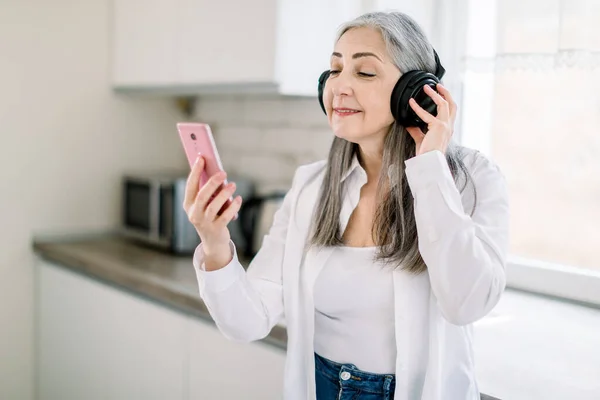 Enjoying music and having fun in the kitchen. Cheerful old gray haired woman is listening to music from earphones and smiling, or havinf video call with best friend. Elderly people and technologies