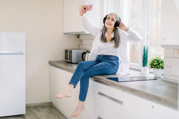 Concepto de gente en casa. Mujer mayor caucásica feliz con auriculares que sostienen el teléfono móvil para hacer una foto o videollamada, mientras está sentado en la encimera de la cocina en la cocina moderna — Foto de Stock