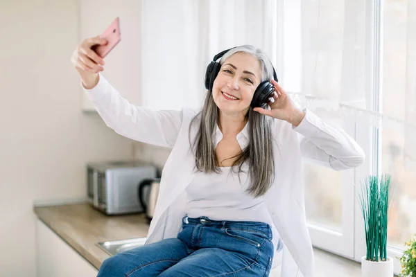 Modern senior people and technologies concept. Smiling retired happy woman using smartphone and headphones for listening music and making video calls or photo, in the kitchen at home. — Stock Photo, Image