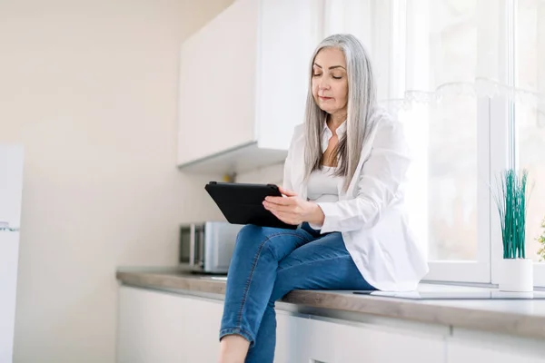 Modern charming senior lady with long gray hair, wearing jeans and white shirt, sitting on the kitchen countertop while surfing social media or cooking recipes on tablet in kitchen