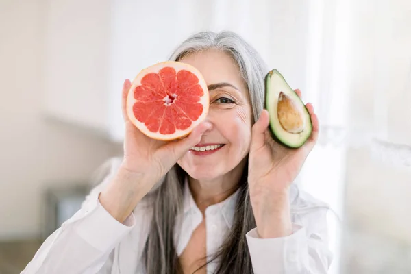 Primer plano retrato de la mujer de pelo gris en la cocina, sosteniendo aguacate fresco y pomelo, sonriendo a cámara oculta su ojo. Alimentación saludable y concepto de comida natural — Foto de Stock