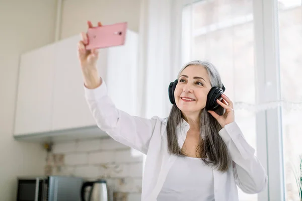 Όμορφη συνταξιούχος γκρίζα μαλλιά γυναίκα σε λευκό πουκάμισο στέκεται στην κουζίνα, ακούγοντας μουσική σε ακουστικά και χρησιμοποιώντας εφαρμογές πολυμέσων στο smartphone της ή κάνοντας selfie φωτογραφία — Φωτογραφία Αρχείου
