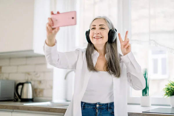 Portrait of positive, happy, smiling senior lady with long gray hair, with headphones, having fun in kitchen at home, listening to music and making photo on cell phone gesturing victory sign — Stock Photo, Image