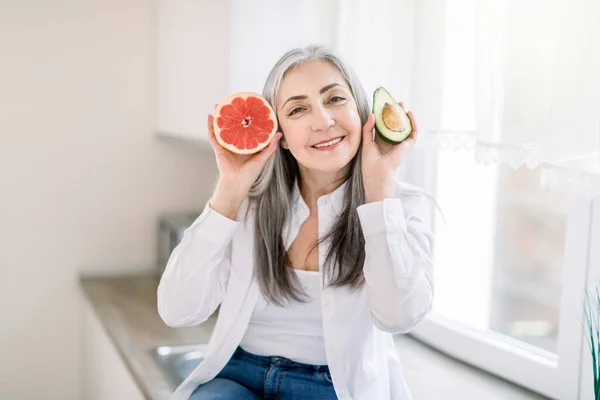 Healthy eating for senior people, vegan food concept. Attractive smiling Caucasian elderly woman sits on the countertop in modern kitchen, holds in hands fresh avocado and grapefruit — Stock Photo, Image