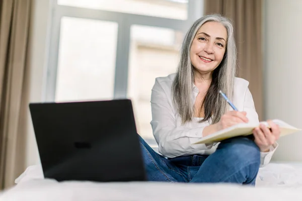 Senior woman with beautiful long gray hair, searching information online on laptop and taking notes in notebook, while sitting in lotus position on bed at home — Stock Photo, Image