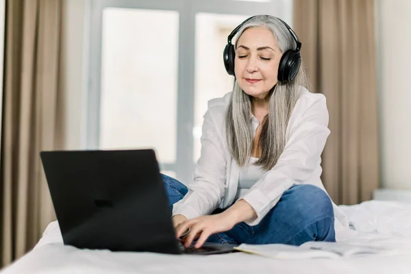 Horizontal shot of smiling gray haired senior woman, sitting on bed in home bedroom, typing on laptop, listening to music or video in headphones and working online or chatting with friend — Stock Photo, Image