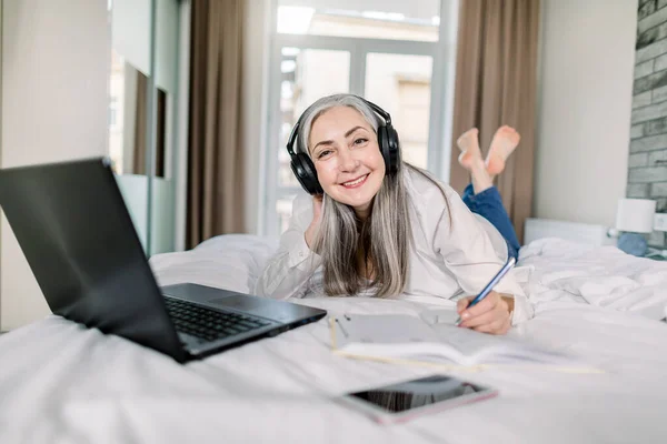 Beautiful senior smiling woman lying down the bed with legs raised slightly, using laptop and making notes in her notepad, working and studying from home. Elderly people and technologies