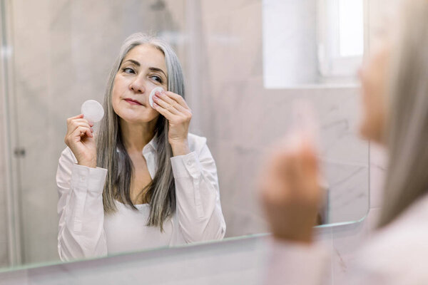 Skin care and hygiene concept. Charming senior gray haired lady using a cotton pad with micellar water for removing make up from face, standing in bathroom and looking in the mirror